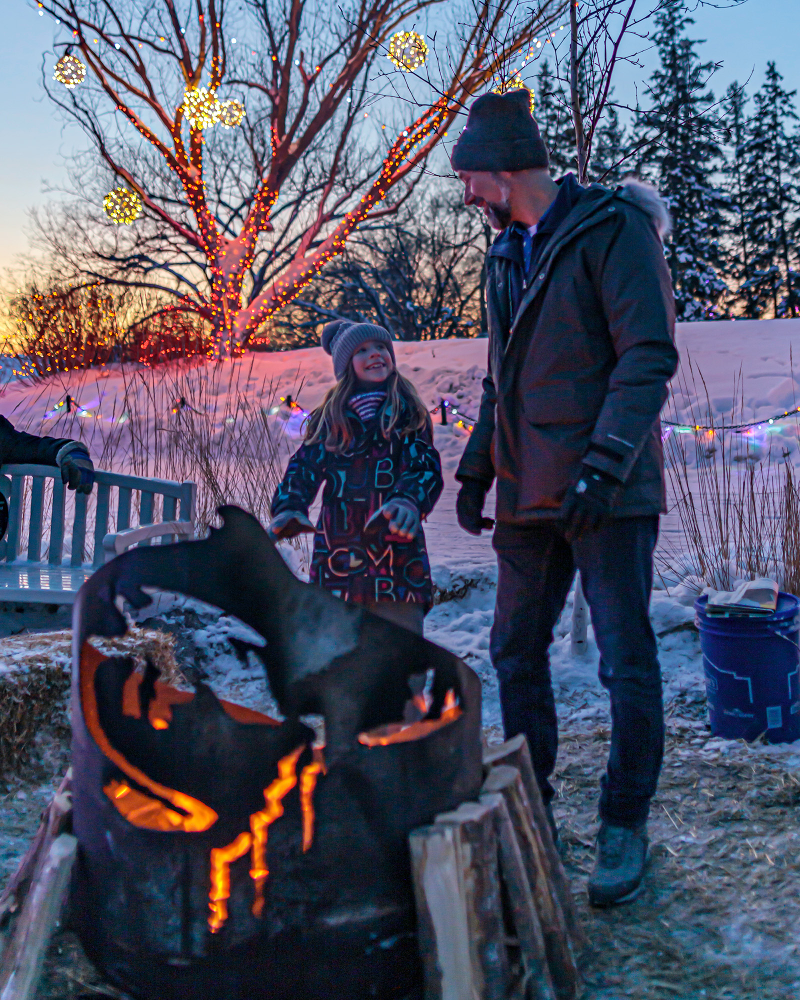 Father and daughter stand behind a fire pit, daughter warms her hands